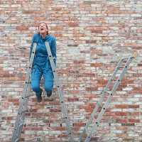 Vor einer alten Backsteinmauer stütz sich eine Person in einem blauen Overall auf zwei Leitern, während die Füße in der Luft baumeln.