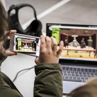 Eine Person filmt mit einem Handy einen Laptopbildschirm ab, auf dem ein Puppenspiel gezeigt wird.