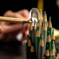 Eine kleine Papierfigur ohne Kopf steht auf mehreren grünen Buntstiften und wird von einem weiteren Stift manuell gestützt.