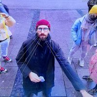 Ein Mann mit einer roten Mütze, einer Brille und Bart wird von einer Kamera gefilmt, die gleichzeitig viele daten über den Mann ausrechnet und auf dem Bildschirm festhält.
