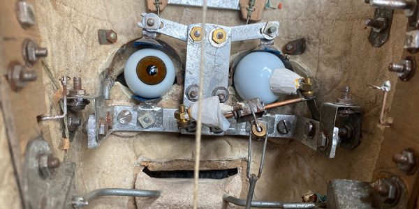 Auf verschiedene Schrauben und Apparaturen ist ein alter Stoff und zwei runde große Augen gespannt. Ein Fertigungsprozess eines Gesichtes.