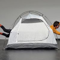 Ein graues Campingzelt vor einer grauen Wand. Aus dem Zelt schauen auf einer Seite Füße, auf der anderen Seite Arme und ein Kopf heraus.
