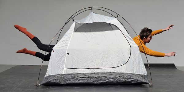 Ein graues Campingzelt vor einer grauen Wand. Aus dem Zelt schauen auf einer Seite Füße, auf der anderen Seite Arme und ein Kopf heraus.