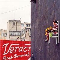 An der Betonwand einer alten heruntergekommenen Fabrik ist ein weißer Stuhl in ungefähr drei Meter Höhe aufgehangen, auf dem eine ältere Dame sitzt, die mit gelben Blumen bastelt. Im Hintergrund sind weitere verfallene Gebäude.