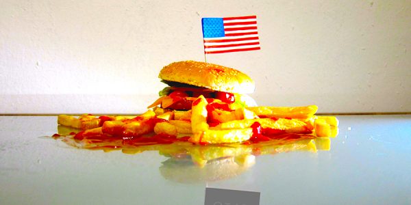 Auf einem Burger mit vielen Pommes und Ketchup steckt eine Amerika Fahne. In der Spiegelung des Tisches, steht statt der Fahne 8:64.