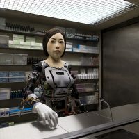 Eine Roboterfrau steht vor einem Tresen. Hinter ihr ist ein Regal mit vielen Boxen und Döschen. Die Frau besteht bis auf ihr Gesicht und ihre Hand nur aus Kabeln und Platten. Das Gesicht und die Hand haben eine Art menschliche Maske
