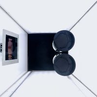 Der Blick geht von oben in eine weiße Box mit quadratischer Grundfläche, in der zwei schwarze Hocker stehen und ein Bildschirm an der Wand hängt.
