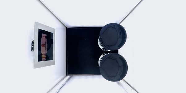 Der Blick geht von oben in eine weiße Box mit quadratischer Grundfläche, in der zwei schwarze Hocker stehen und ein Bildschirm an der Wand hängt.